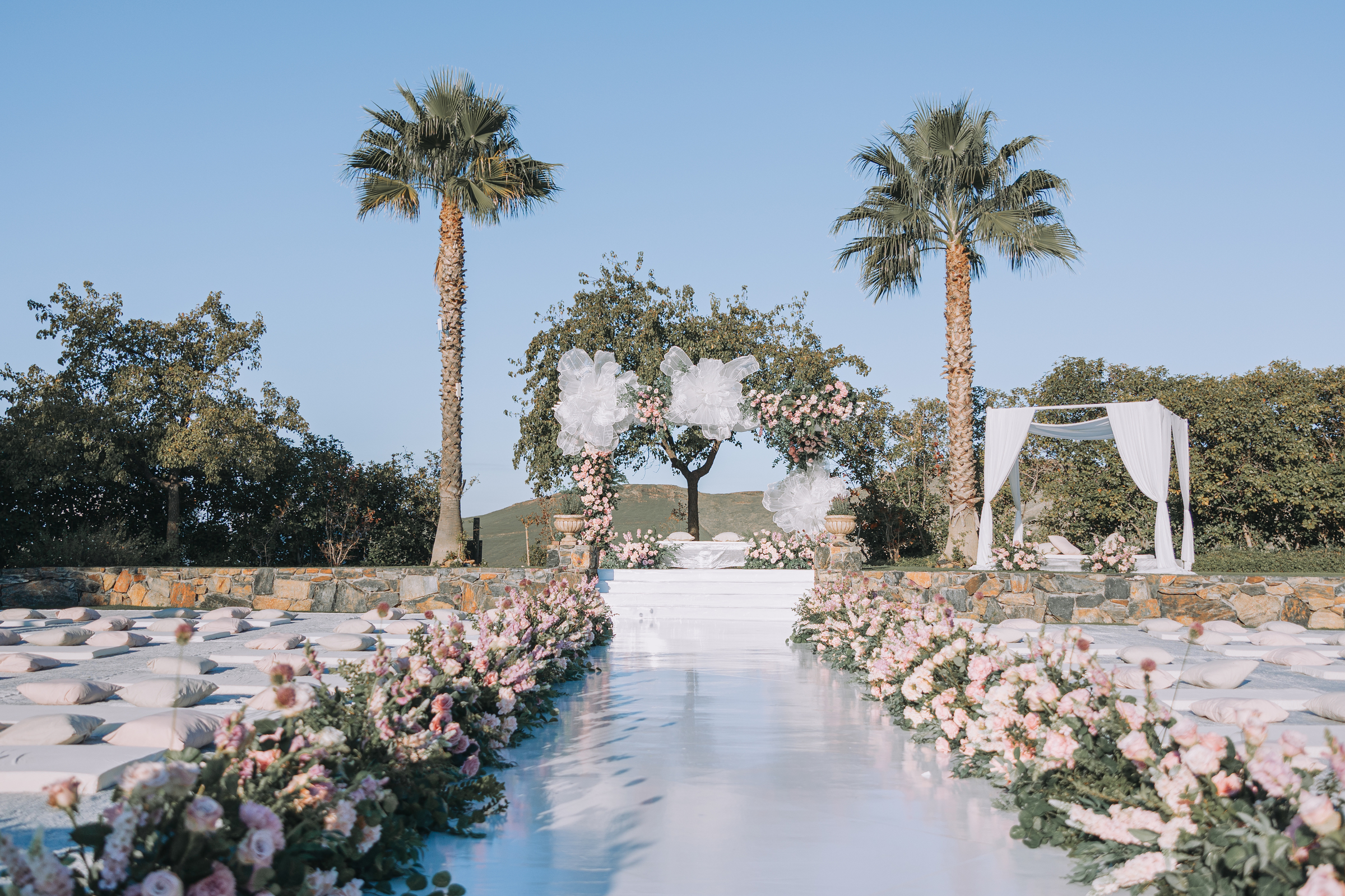 A ceremony aisle lined with roses leading to a floral arch, framed by palms above the foothills