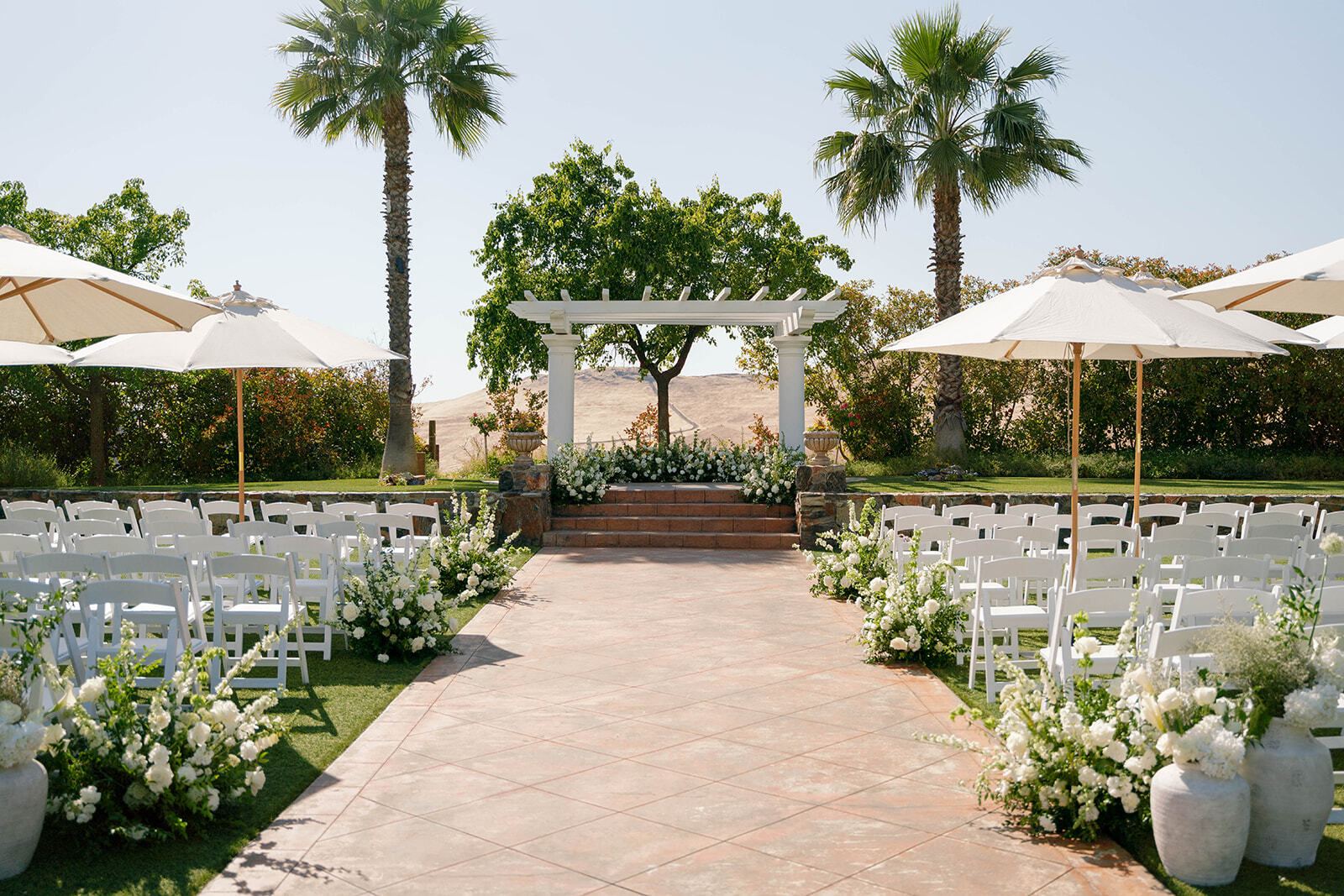 A ceremony set at the Garden Pavilion — white chairs and florals beneath the columned pergola, foothills behind